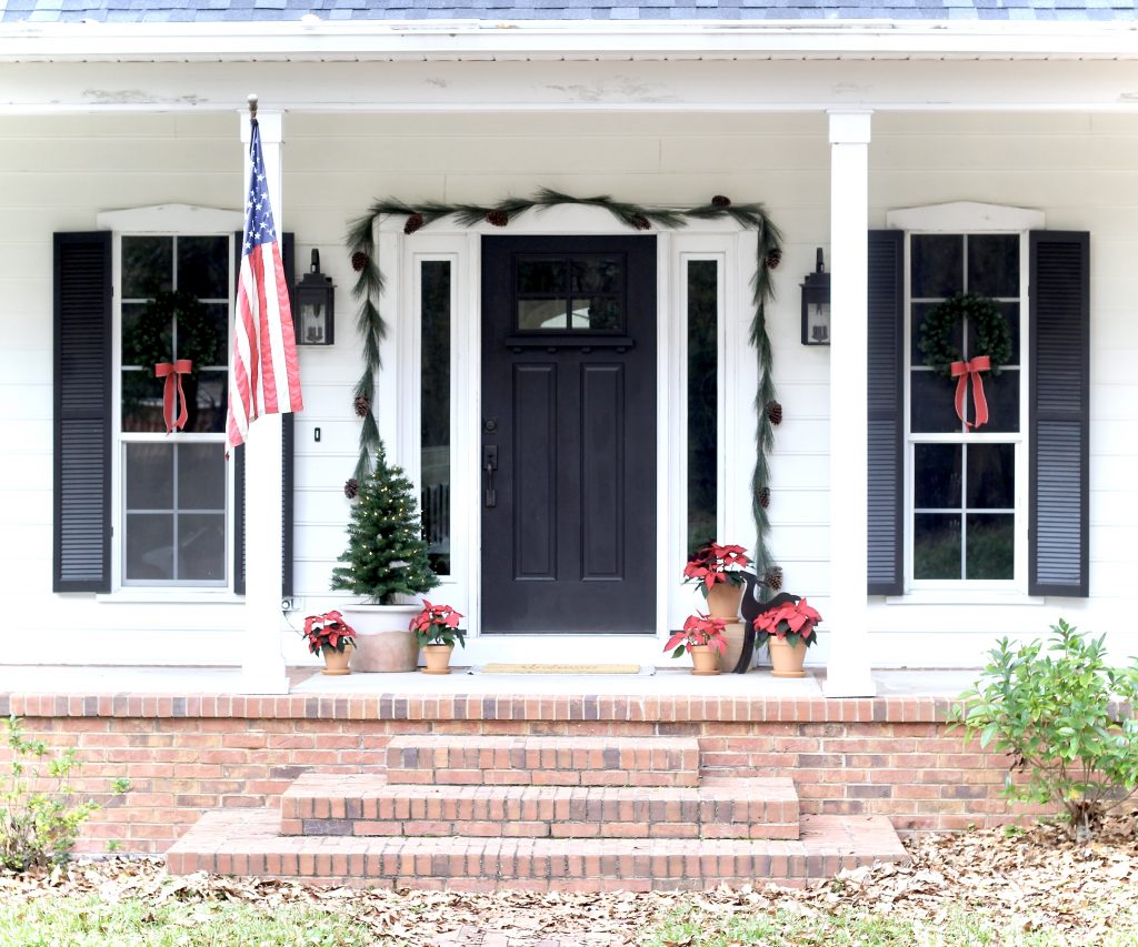White modern farmhouse with black shutters, wreaths with red bows on the windows, and garland over the front door| #modernfarmhouse #frontporch #Christmasdecor #wreath