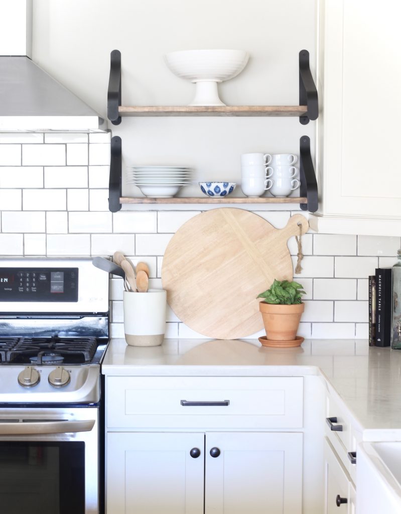 Open shelving in white kitchen with round cutting board, white dishes, and plant. #whitekitchen #openshelving #shelfie #cottagedecor 