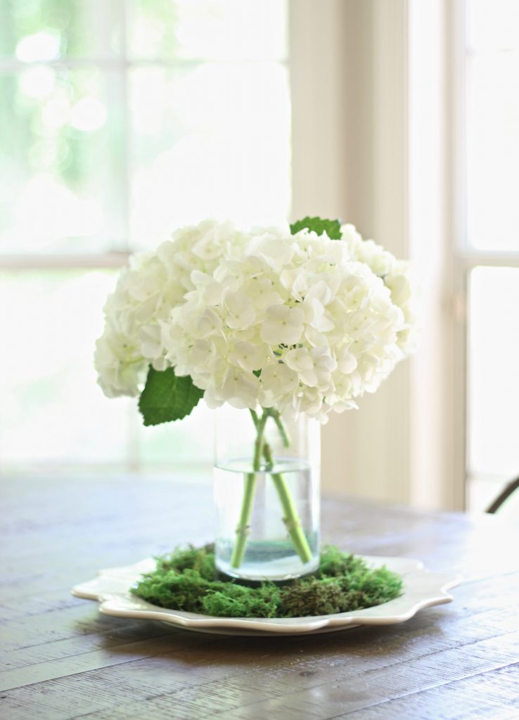 Simple floral centerpiece of white hydrangeas in glass vase surrounded by green moss | www.ourhammockhouse.com | #whitehydrangea #centerpiece #springdecor #summerdecor 